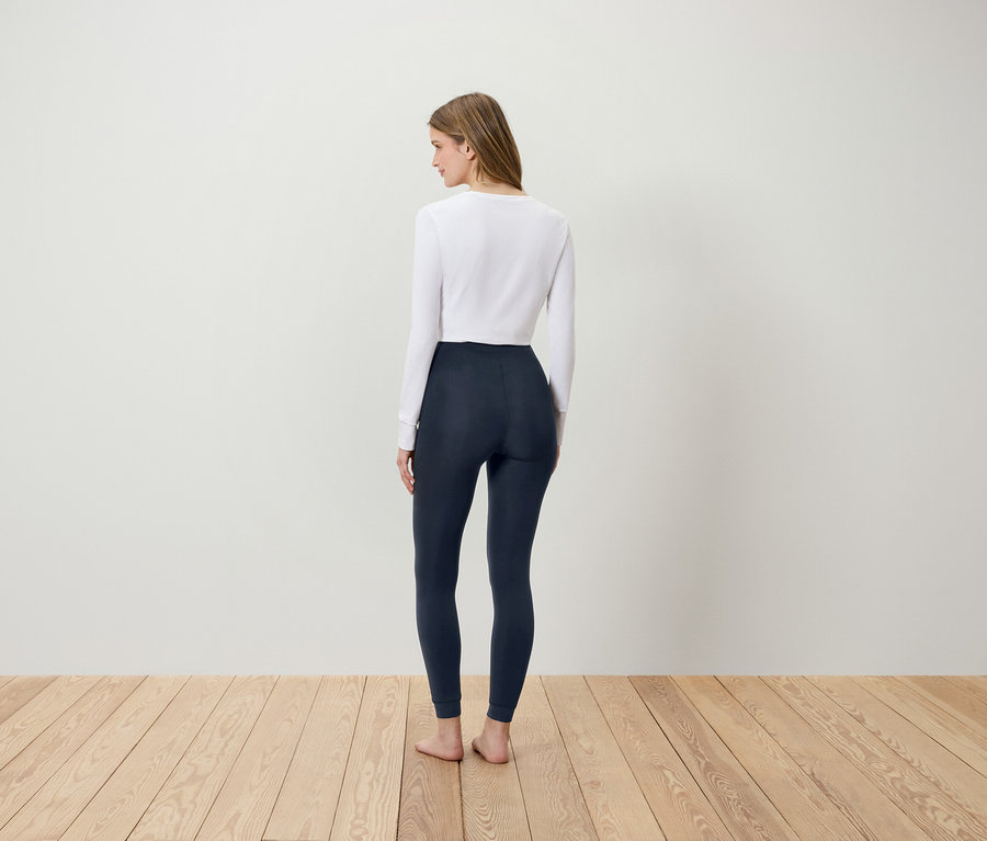 Frau in weißem Shirt und dunkelblauen Leggings steht mit dem Rücken zum Betrachter auf einem Holzboden.