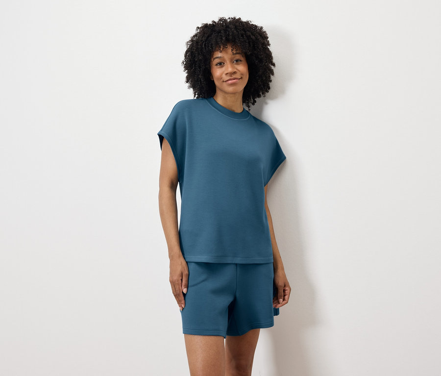 Frau mit lockigem Haar, in blauem T-Shirt und Shorts, steht vor einer weißen Wand.