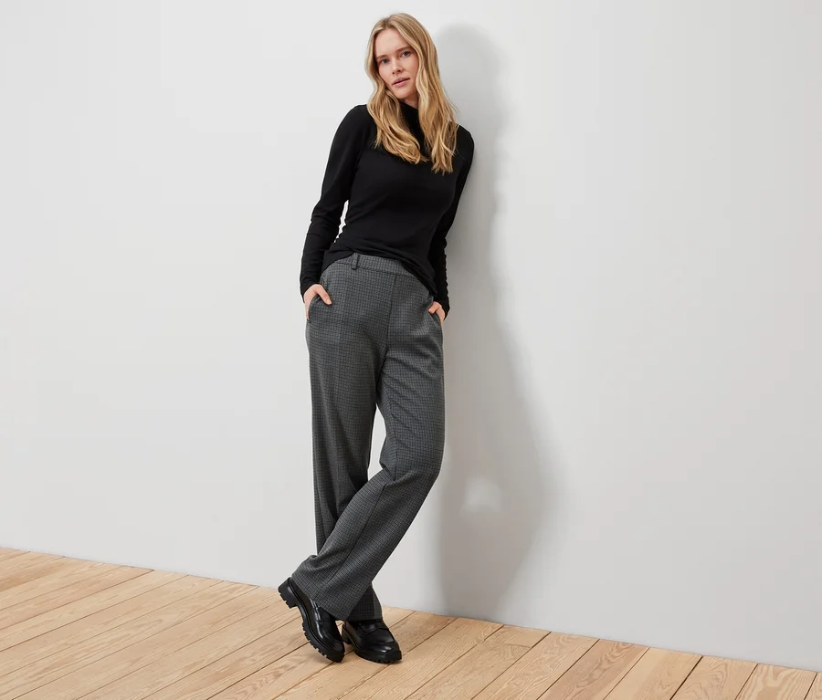 Blonde Frau posiert in schwarzem Rollkragenpullover, grauer Karohose und schwarzen Schuhen.