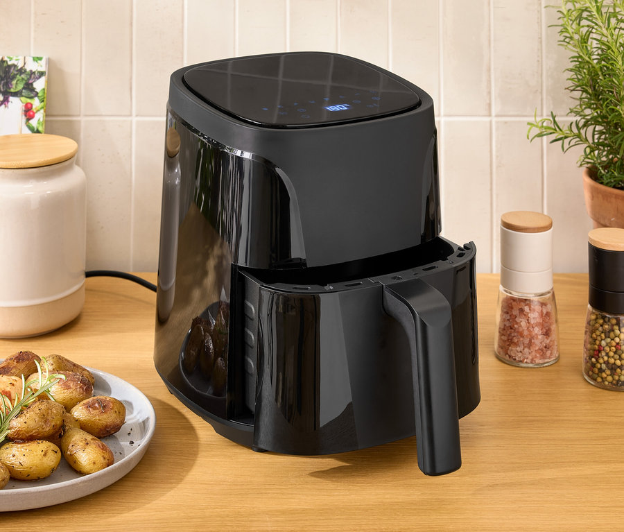 Eine schwarze Heißluftfritteuse steht auf einem Holztisch neben einem Teller mit Hühnchen und Kartoffeln.