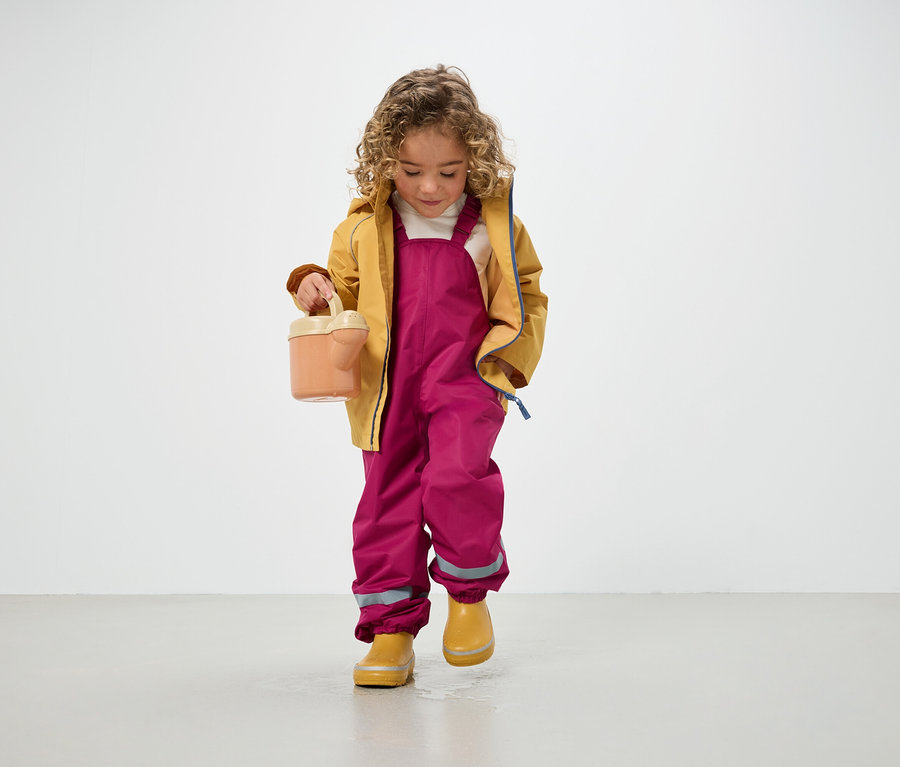 Ein lockiges Kind in gelber Jacke, rosafarbenen Hosen und gelben Gummistiefeln hält eine Gießkanne und steht auf dem Boden.