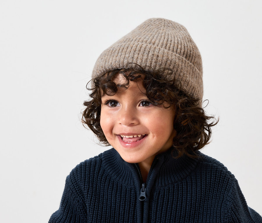 Lächelnder Junge mit braunen lockigen Haaren, der eine beige Wollmütze und eine dunkelblaue Strickjacke mit Reißverschluss trägt.