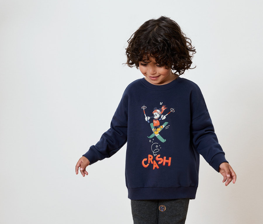 Ein Kind mit dunklem lockigem Haar trägt ein dunkelblaues Sweatshirt mit einem Bild von Mickey Mouse beim Skifahren.