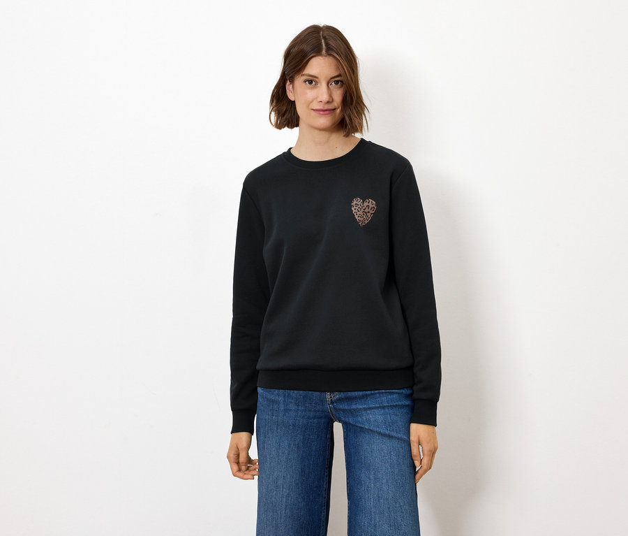 Frau steht und trägt einen schwarzen Pullover mit Herzapplikation und Jeans.