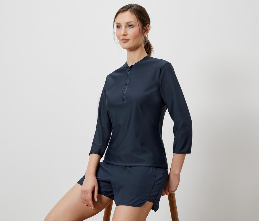 Frau sitzt auf einem Hocker und trägt ein UV-Schutz-Sportshirt und Shorts.