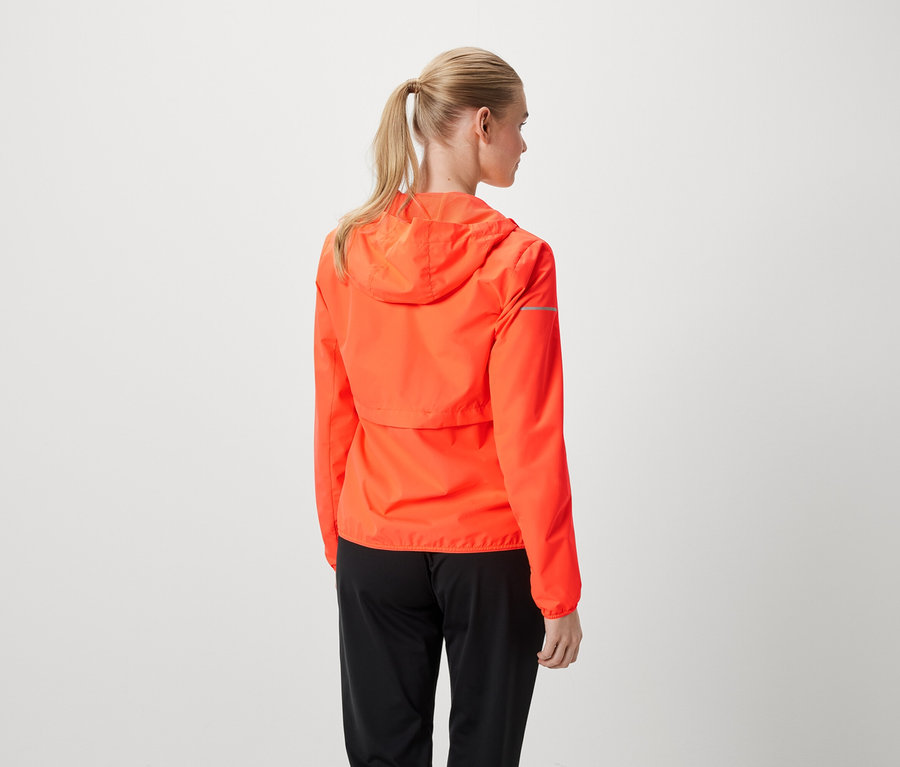 Frau mit blonden Haaren im Pferdeschwanz trägt eine orangefarbene Jacke und schwarze Hose.