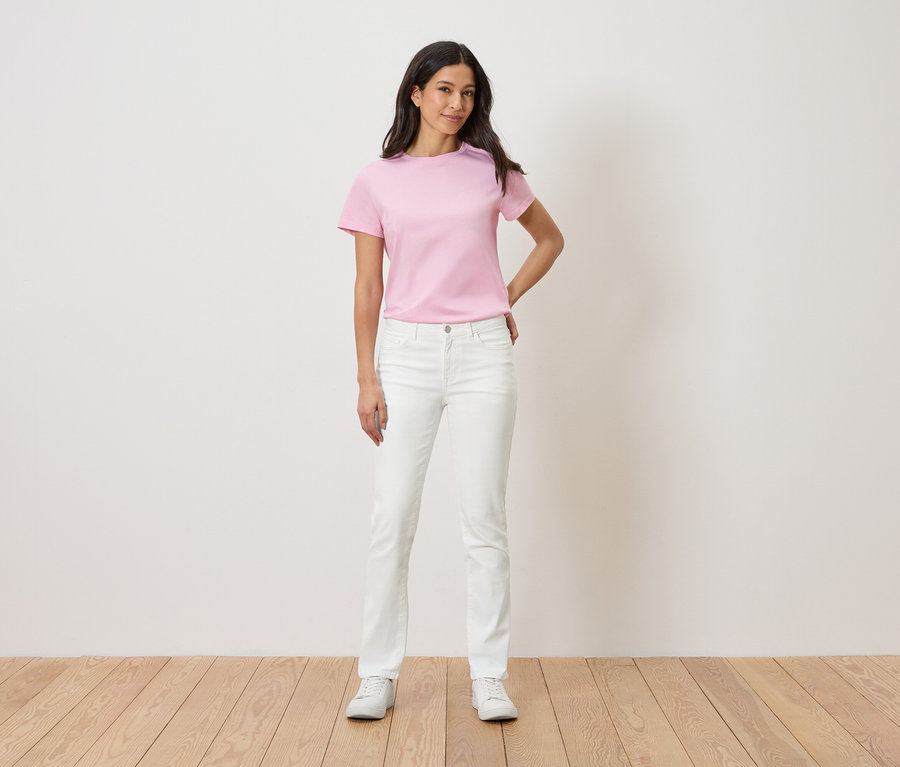 Frau steht vor einer weißen Wand und trägt ein rosa T-Shirt, weiße Jeans und weiße Turnschuhe.