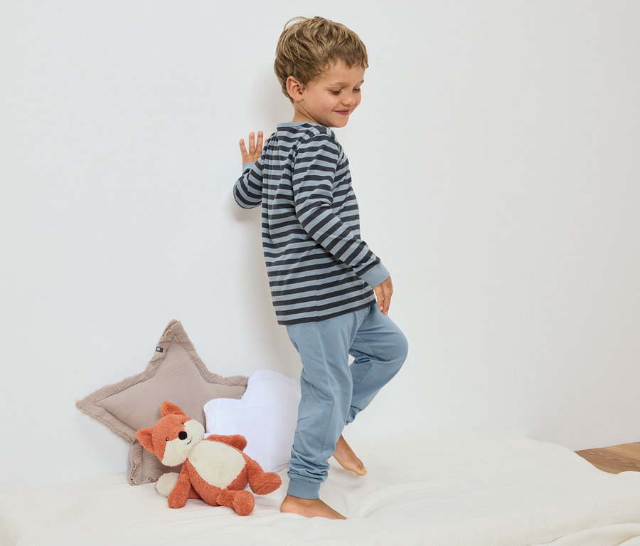 Ein kleiner Junge in gestreiftem Schlafanzug steht auf einem Bett neben einem Fuchs-Stofftier und sternförmigen Kissen.