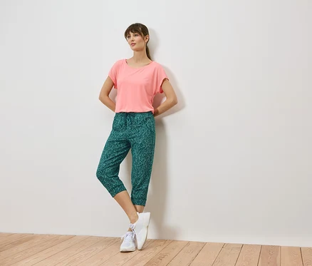 Frau lehnt an einer Wand, trägt ein korallfarbenes T-Shirt, grüne Caprihosen mit Blattmuster und weiße Turnschuhe.