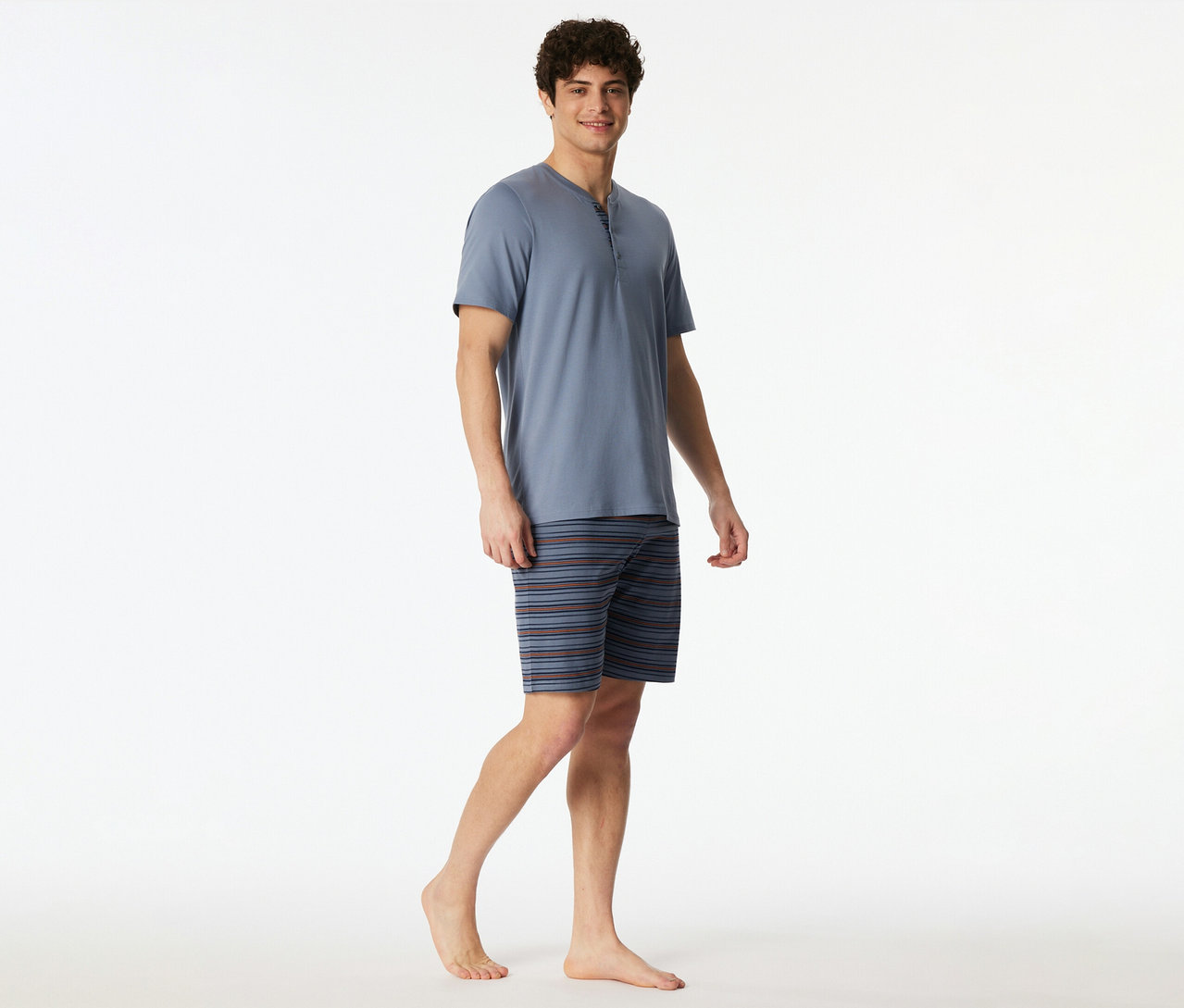 Ein lächelnder Mann trägt ein blaues SCHIESSER Shorty-Set mit Kurzarm-Henley-Oberteil und gestreiften Shorts, barfuß stehend.