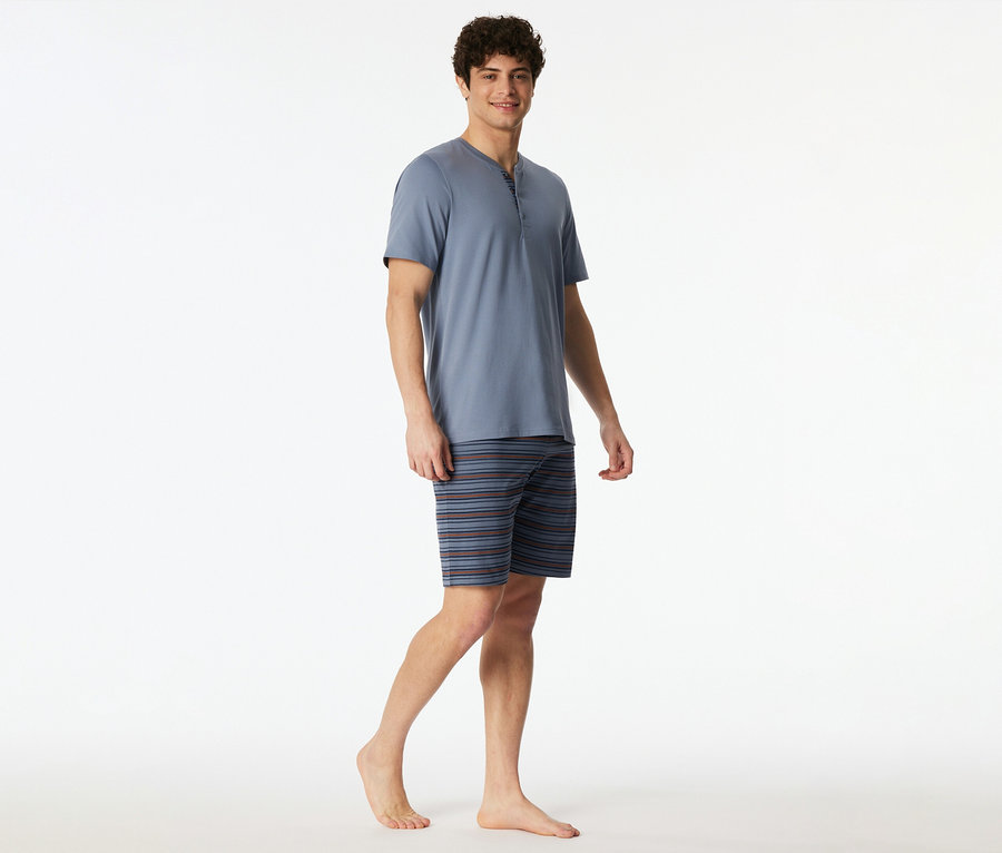 Ein lächelnder Mann trägt ein blaues SCHIESSER Shorty-Set mit Kurzarm-Henley-Oberteil und gestreiften Shorts, barfuß stehend.