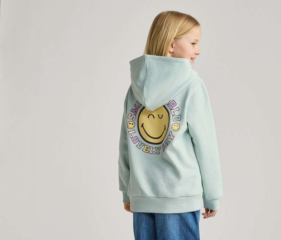 Ein Mädchen steht mit dem Rücken zur Kamera und trägt einen hellblauen Hoodie mit einem Smiley-Aufdruck.