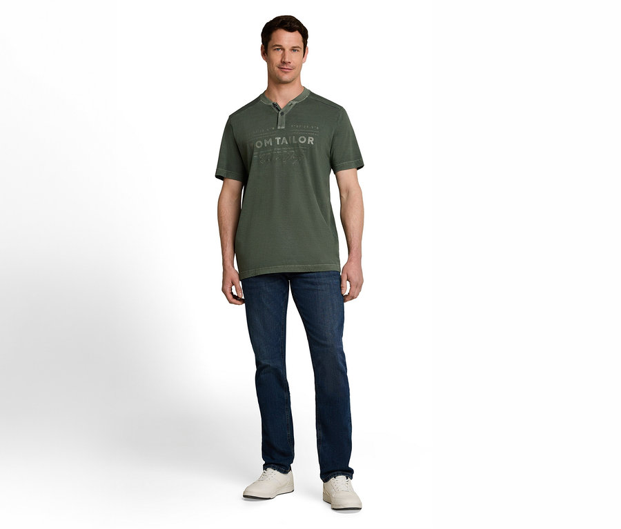 Ein Mann steht frontal, trägt ein dunkelgrünes kurzärmeliges Henley-Shirt mit dezentem Aufdruck, dunkelblaue Jeans und weiße Turnschuhe.