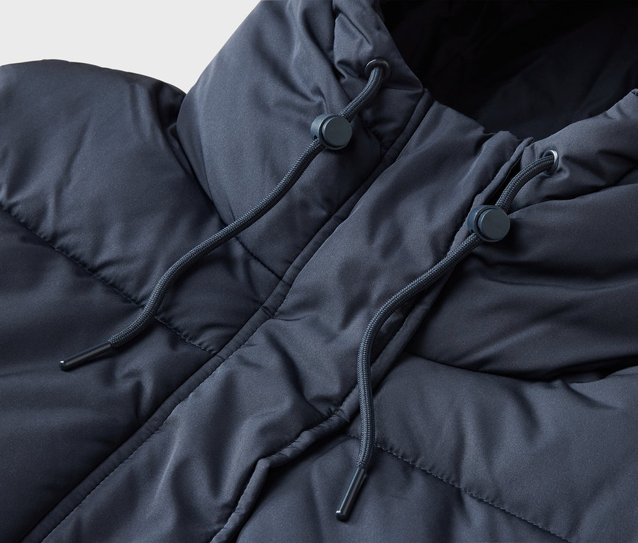 Nahaufnahme einer dunkelblauen Steppjacke mit Kapuze, die zwei Kordelzüge mit dunkelblauen Stoppern zeigt. Die Jacke hat eine gesteppte, bauschige Textur.