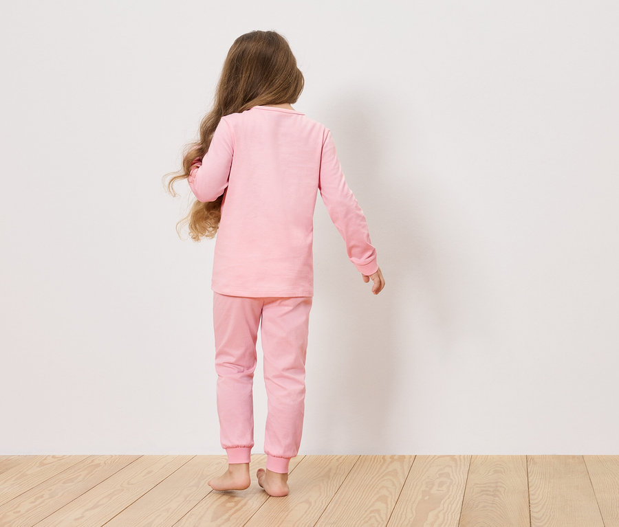 Mädchen mit langen braunen Haaren, von hinten gesehen, trägt einen rosa Pyjama vor einer weißen Wand.