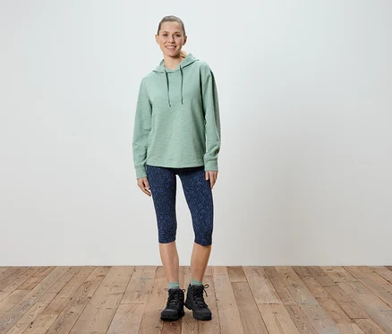 Frau steht in hellgrünem Hoodie, gemusterten blauen Capri-Leggings und schwarzen Wanderschuhen.