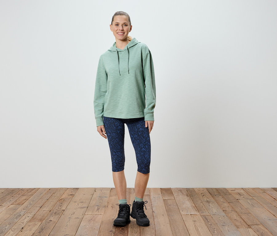 Frau steht in hellgrünem Hoodie, gemusterten blauen Capri-Leggings und schwarzen Wanderschuhen.