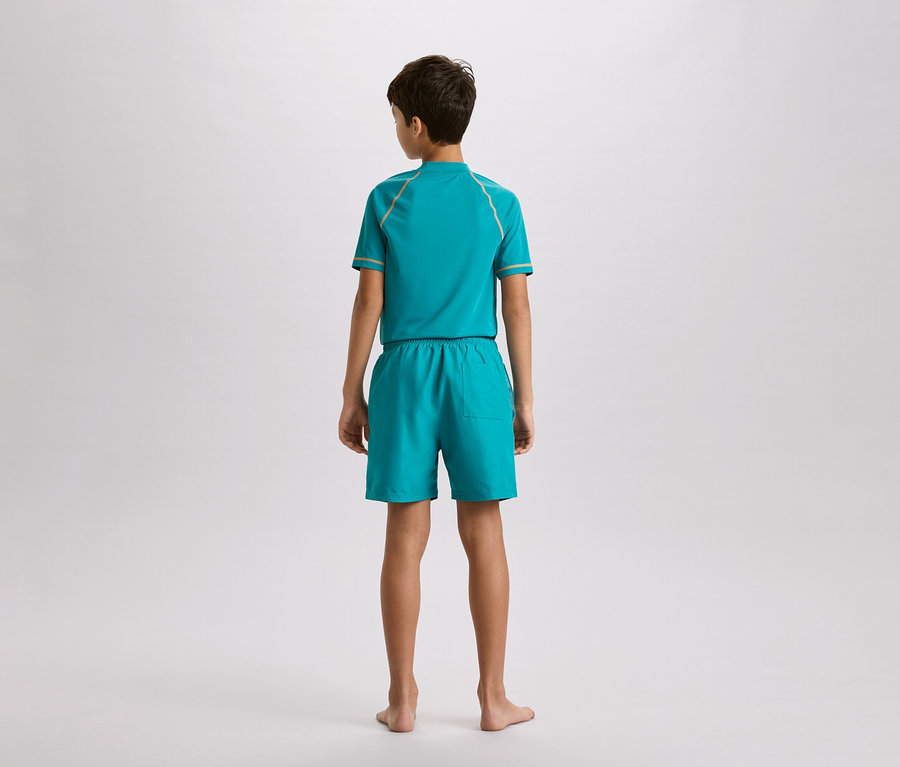 Junge von hinten, trägt ein türkisfarbenes Set aus Oberteil und Shorts mit goldfarbenen Akzenten, vor einem neutralen Hintergrund.