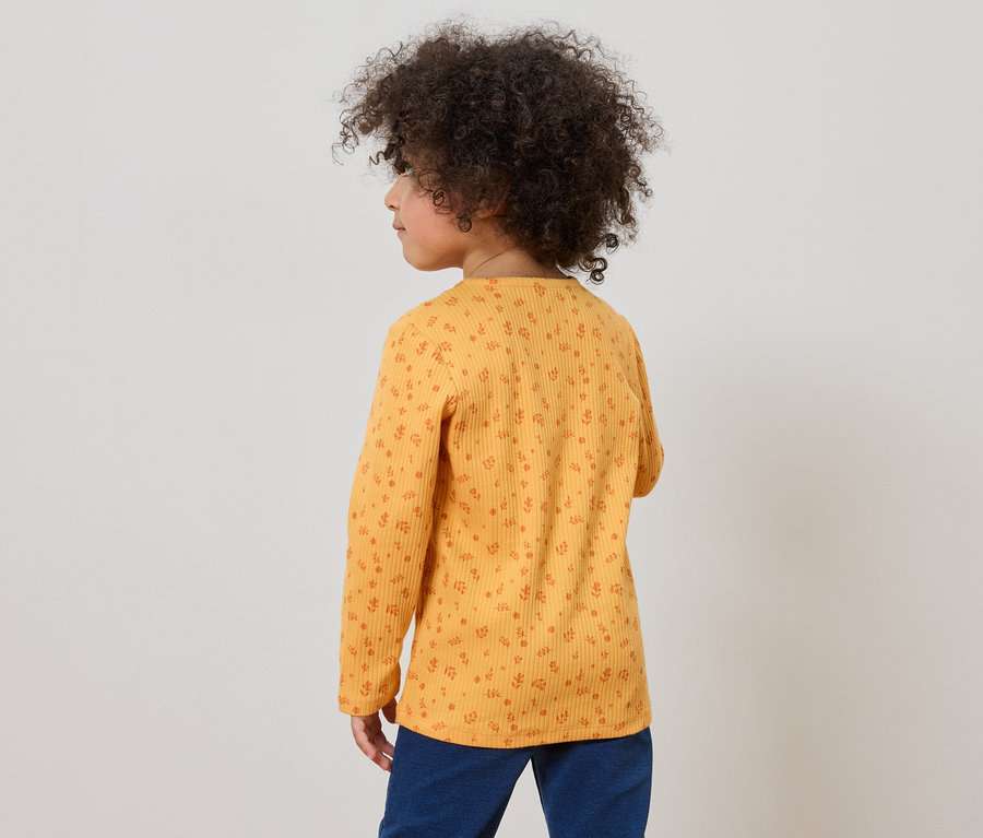 Kind mit lockigem Haar trägt ein gelbes, langärmeliges Oberteil mit Blumenmuster und blaue Hosen, von hinten gesehen.