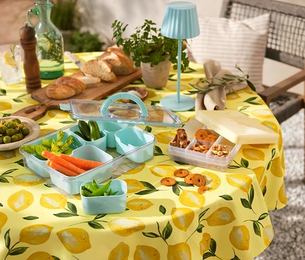 Gelbe, abwischbare Tischdecke mit Zitrone unter Snacktray To-Go mit Gemüse und Keksen, kleiner LED-Tischleuchte, Brot, Getränken und Oliven.