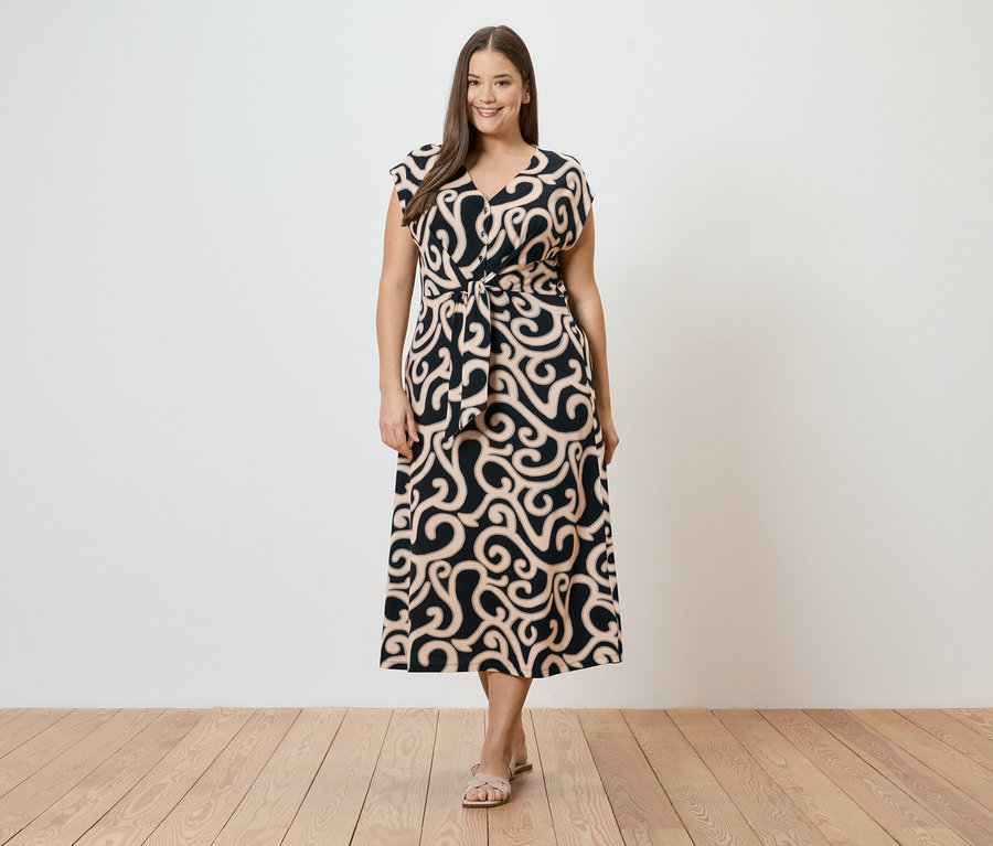 Eine Frau steht vor einer weißen Wand und trägt ein schwarzes, gemustertes Kleid mit Gürtel und Sandalen.