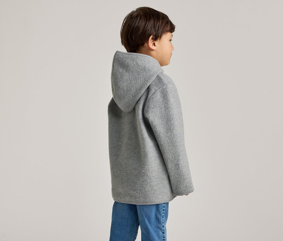 Ein Kind mit braunen Haaren trägt eine graue Kapuzenjacke und Jeans, von der Seite gesehen.
