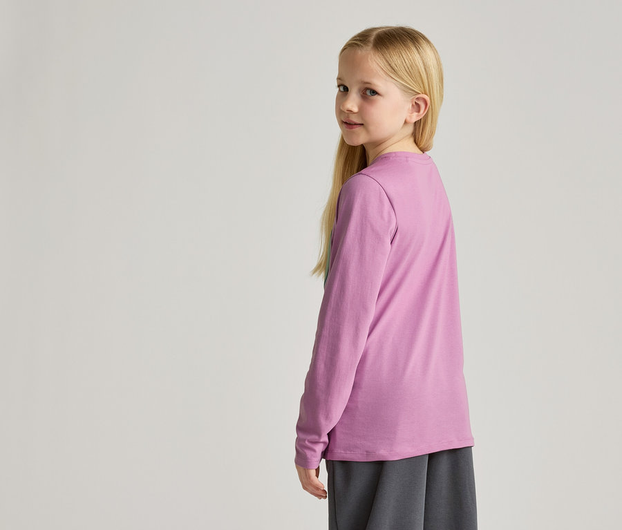 Ein junges Mädchen mit blonden Haaren trägt ein lila Langarmshirt und eine graue Hose.