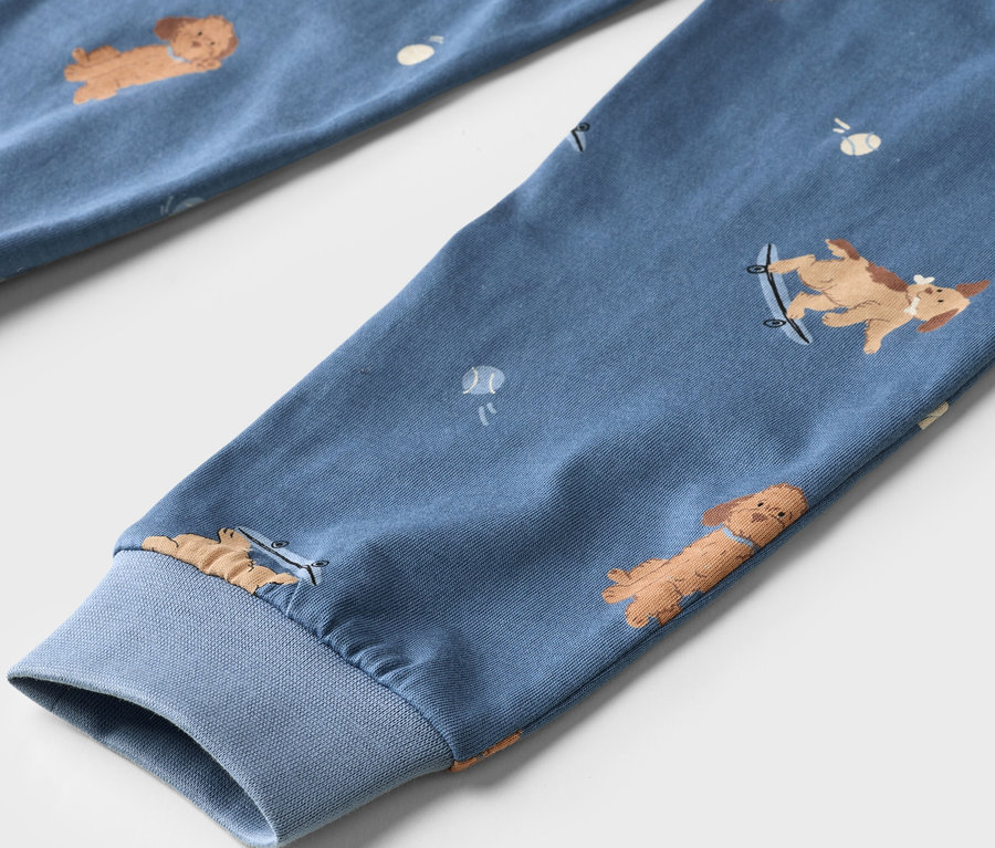 Nahaufnahme eines blauen Stoffes mit einem Muster aus braunen Cartoon-Hunden und weißen Tennisbällen. Ein Hund ist auf einem Skateboard. Der Stoff hat ein geripptes Bündchen.