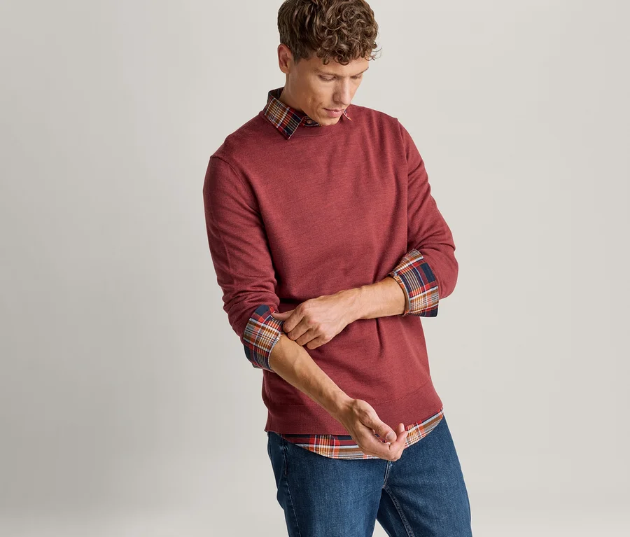 Mann krempelt die Ärmel seines roten Pullovers hoch, unter dem ein kariertes Hemd sichtbar wird, und trägt blaue Jeans.