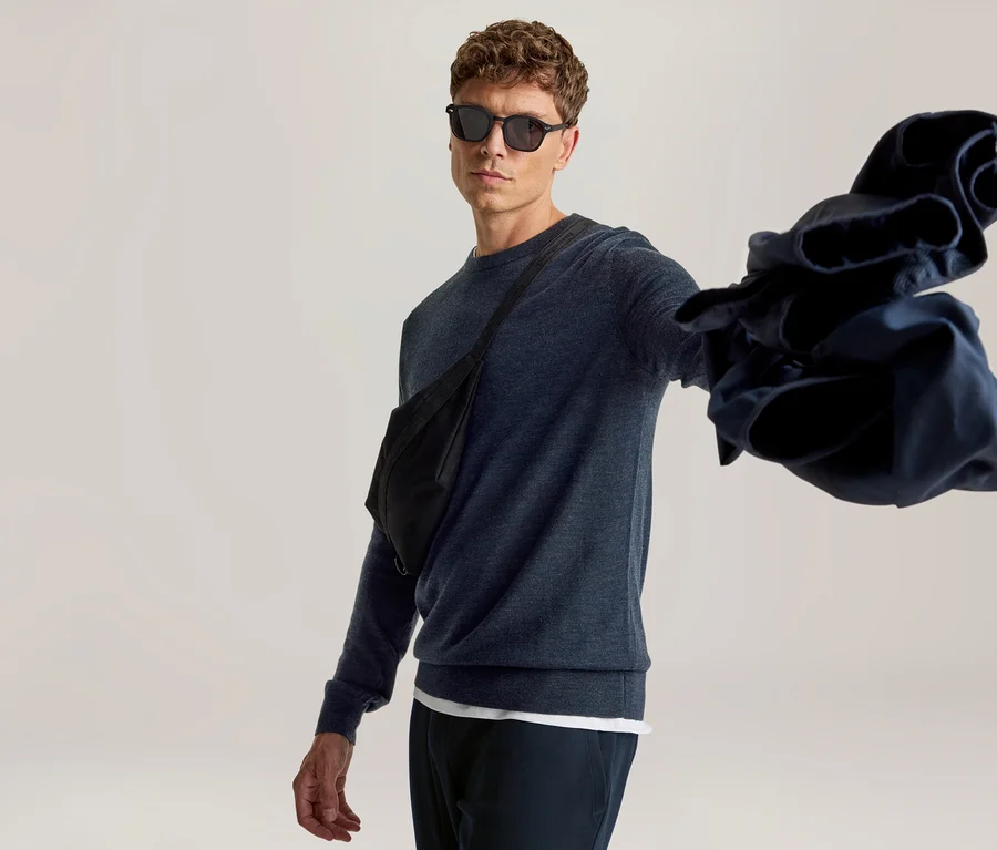 Mann mit Sonnenbrille, dunkelblauem Pullover und schwarzer Bauchtasche, der ein zerknülltes dunkelblaues Kleidungsstück in seiner ausgestreckten Hand hält.