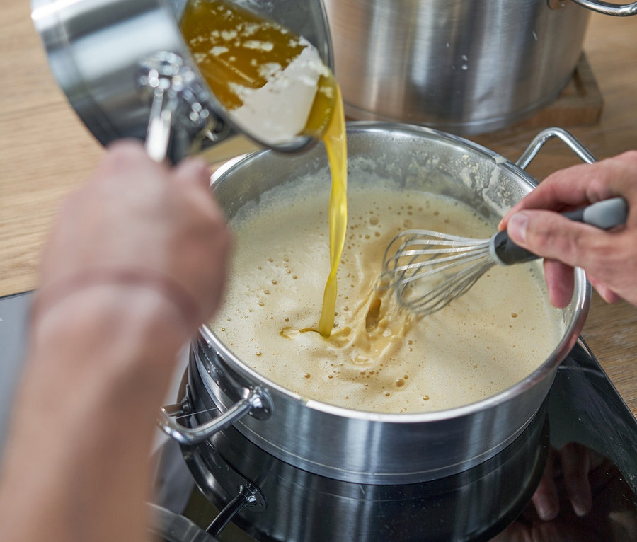 Eine Person gießt geschmolzene Butter aus einem kleinen Topf in einen größeren Topf mit einer hellen, schaumigen Masse und rührt gleichzeitig mit einem Schneebesen um. Im Hintergrund steht ein Dampfgarer.