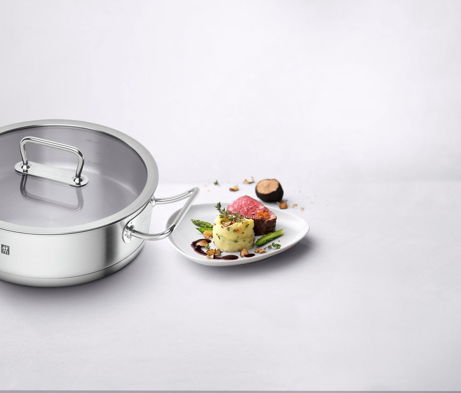 Ein Edelstahltopf mit Glasdeckel steht links neben einem weißen Teller mit einem Gericht aus geschnittenem, rosa Fleisch, Kartoffelpüree und grünem Spargel.