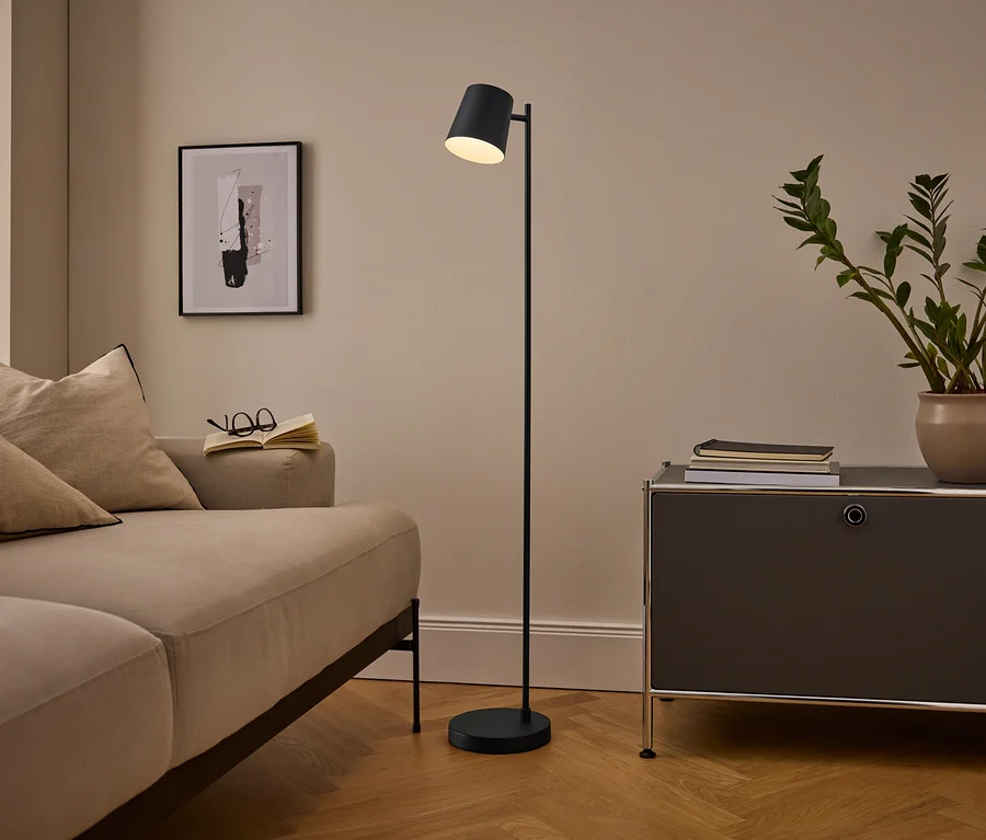 Gemütliches Wohnzimmer mit beigem Sofa, schwarzer Stehlampe und dunkelgrauem Schrank. Ein offenes Buch und eine Brille liegen auf dem Sofa, eine Topfpflanze und Bücher auf dem Schrank. Ein gerahmtes abstraktes Bild hängt an der Wand.