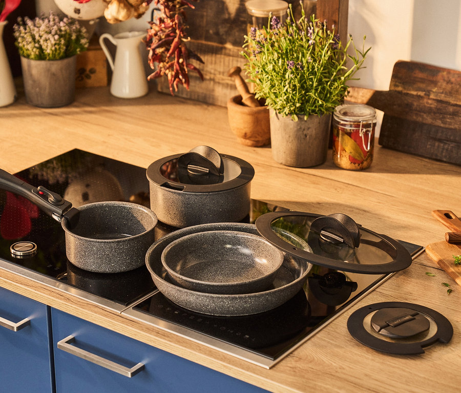 Eine Küchenarbeitsplatte aus Holz mit einem grauen, gesprenkelten Topfset auf einem schwarzen Induktionskochfeld. Im Hintergrund stehen Pflanzen und Küchenutensilien.