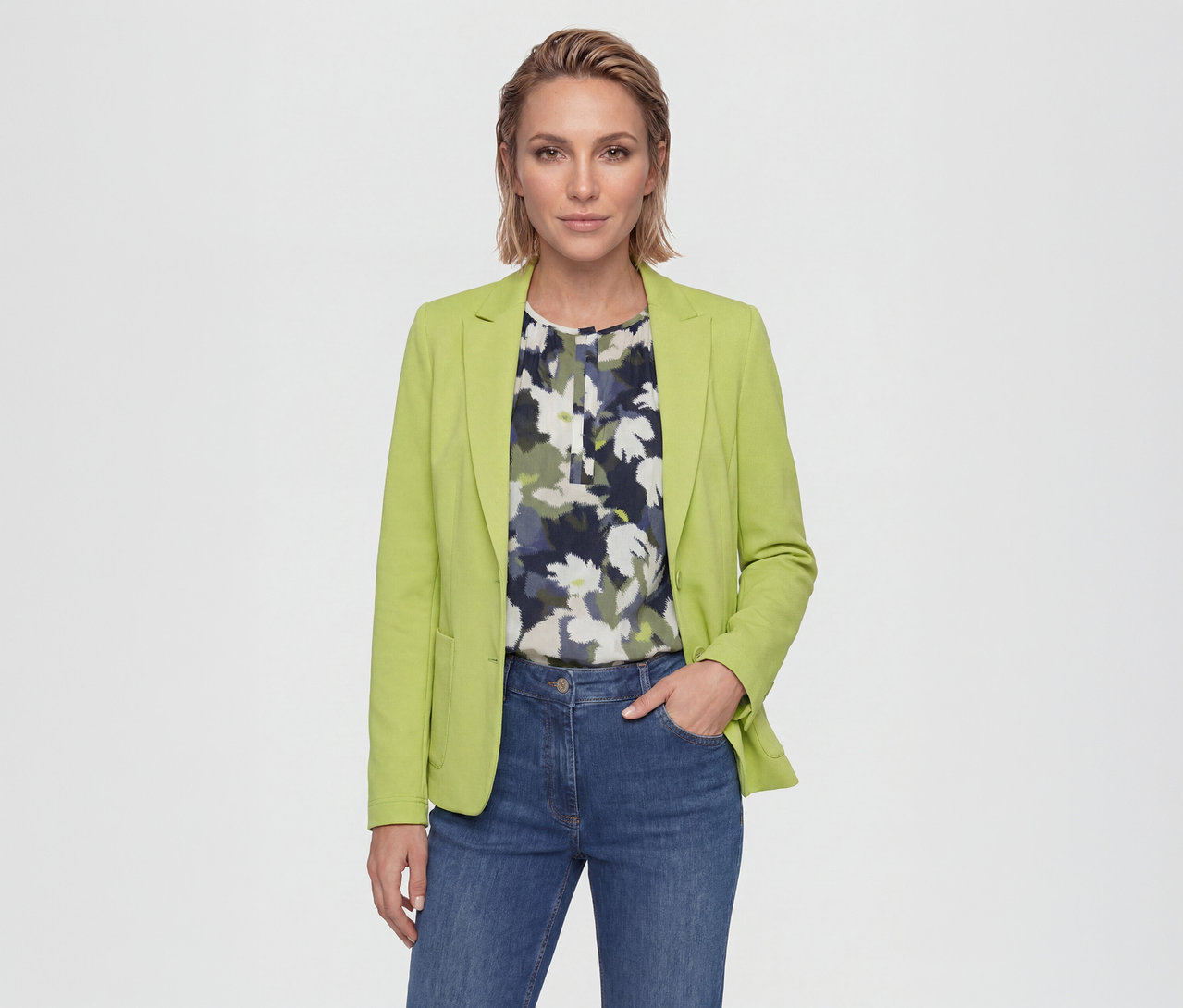 Frau trägt einen limettengrünen Blazer über einer floralen Bluse und blauen Jeans, steht mit einer Hand in der Tasche und blickt in die Kamera.