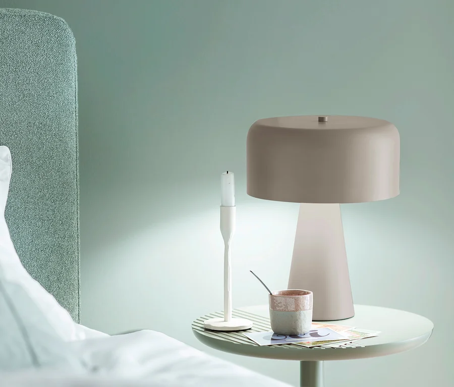 Nachttisch mit einer SCHÖNER WOHNEN-Kollektion Tischleuchte „Pento“ in Sandbrise, einem weißen Kerzenständer und einer Tasse neben einem Bett mit grünem Kopfteil.
