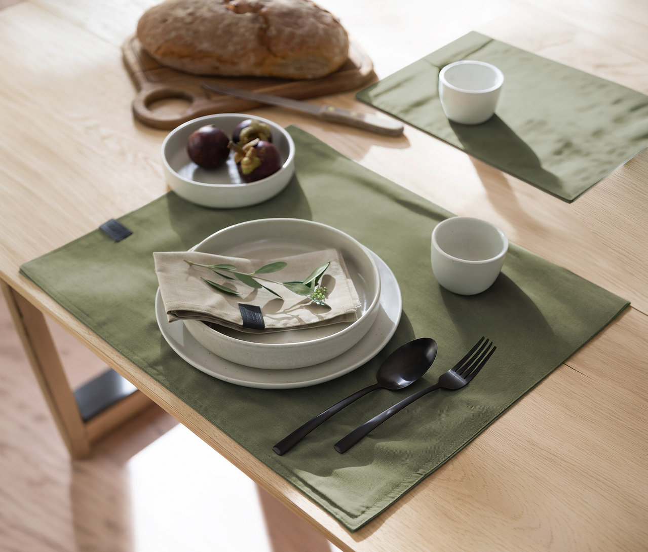 Ein Holztisch ist mit zwei khakifarbenen SCHÖNER WOHNEN-Kollektion Tischsets gedeckt. Darauf stehen weiße Teller mit Serviette und grünem Zweig, schwarzes Besteck, zwei weiße Tassen. Im Hintergrund Brot, Messer und Mangostanen.