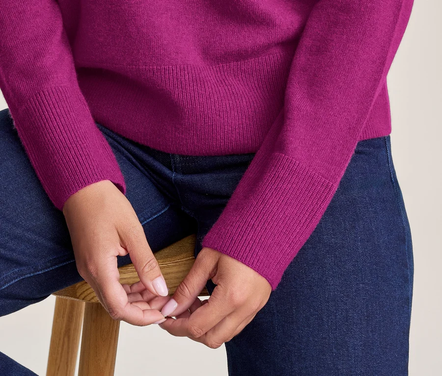 Eine Person in einem fuchsiafarbenen Pullover und dunklen Jeans sitzt auf einem Holzschemel mit verschränkten Händen.