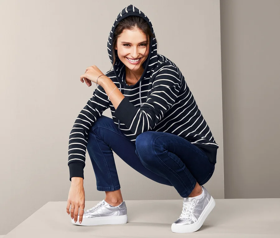 Frau in gestreiftem Kapuzensweatshirt und Jeans posiert auf dem Boden.