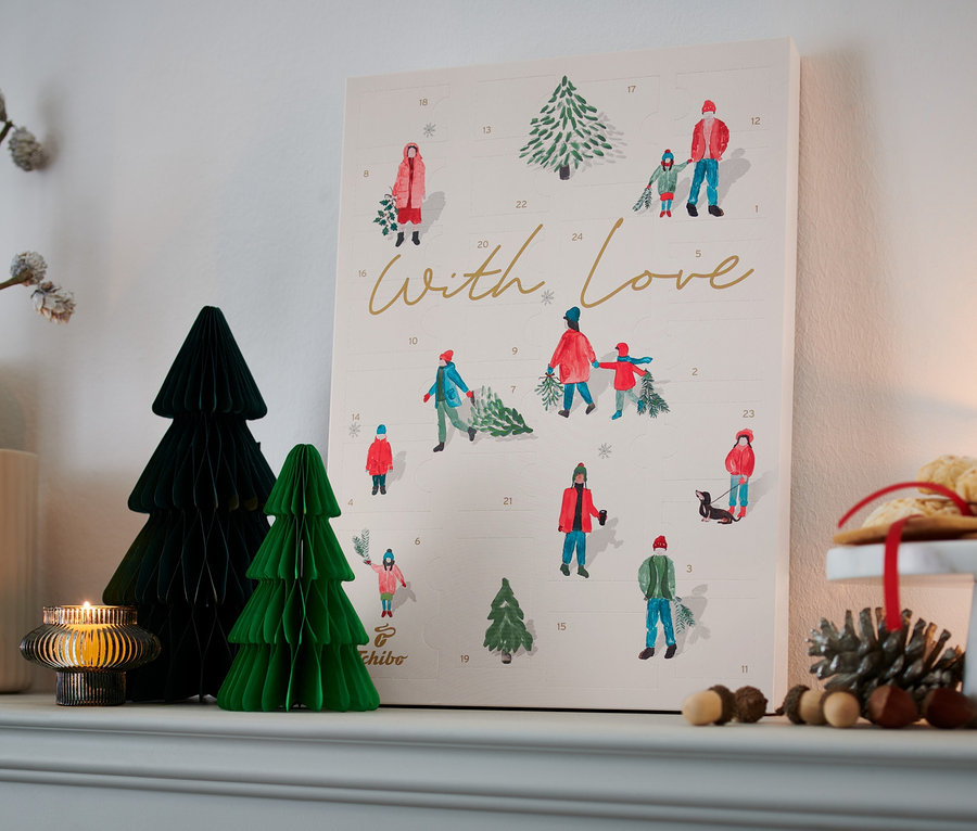 Auf einem Regal stehen Dekorationsartikel, darunter ein Tchibo Adventskalender, eine Vase mit Zweigen, Papierbäume und eine Kerze.