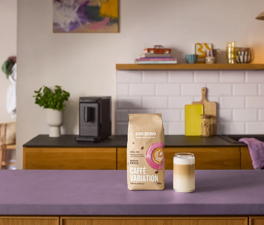 Ein brauner Beutel Eduscho Caffè Variation Ganze Bohne und ein geschichtetes Kaffeegetränk in einem Glas stehen auf einer lila Arbeitsplatte. Im Hintergrund steht ein schwarzer »Esperto mini« Kaffeevollautomat auf einer Küchenzeile mit Spüle und Dekoartikeln.