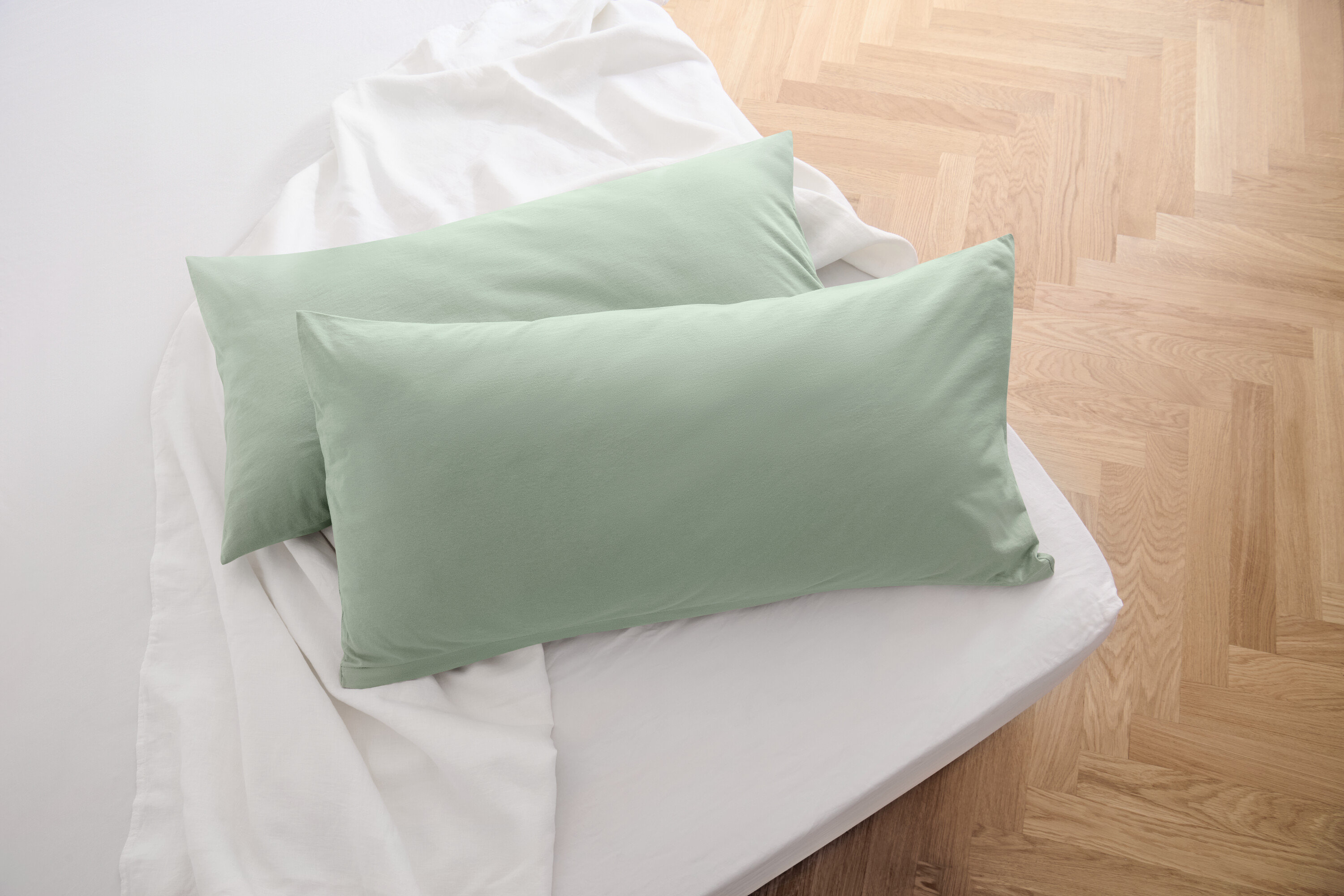 Zwei salbeigrüne Jersey-Kopfkissenbezüge liegen auf einem weißen Bettlaken.