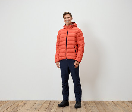 Ein Mann steht und trägt eine orangefarbene Daunenjacke, marineblaue Hosen und schwarze Schuhe.