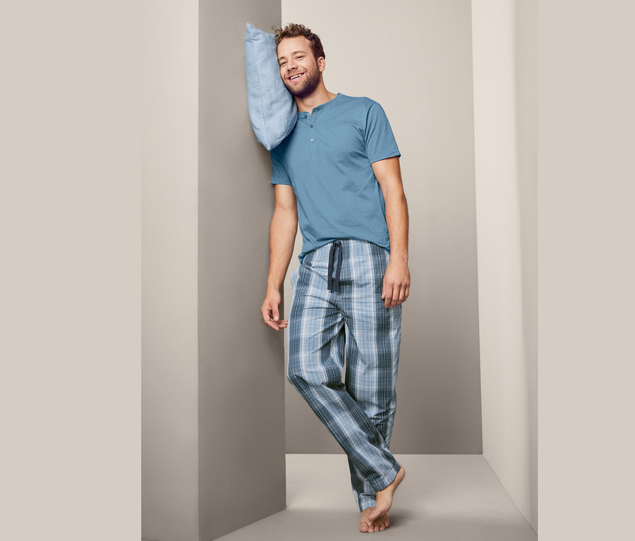 Mann lehnt an einer Wand und hält ein hellblaues Kissen an seinem Kopf. Er trägt einen blauen Pyjama und karierte Hosen.