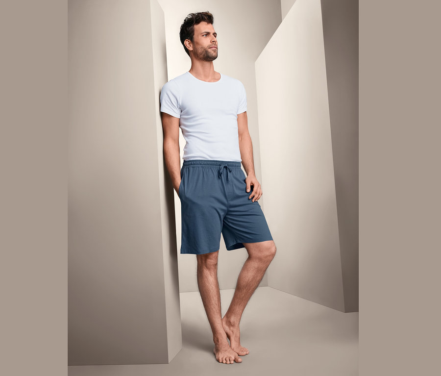 Mann lehnt an Wand, trägt T-Shirt und blaue Jersey-Lounge-und-Schlafshorts.