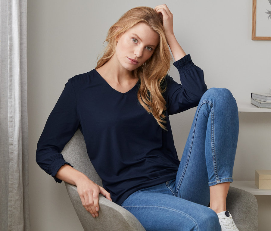 Frau sitzt im Sessel und trägt ein dunkelblaues Blusenshirt mit 3/4-Arm.