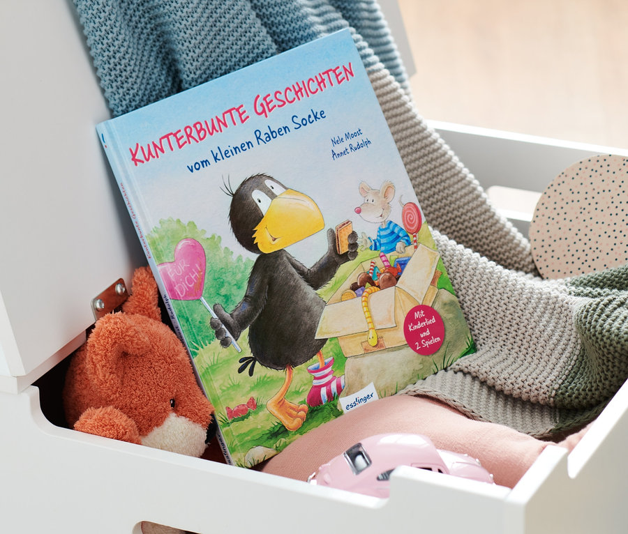 In einer weißen Truhe liegt das Buch »Kunterbunte Geschichten vom kleinen Raben Socke« zusammen mit einem Plüschtierfuchs, einem rosa Spielzeugauto und Strickdecken.