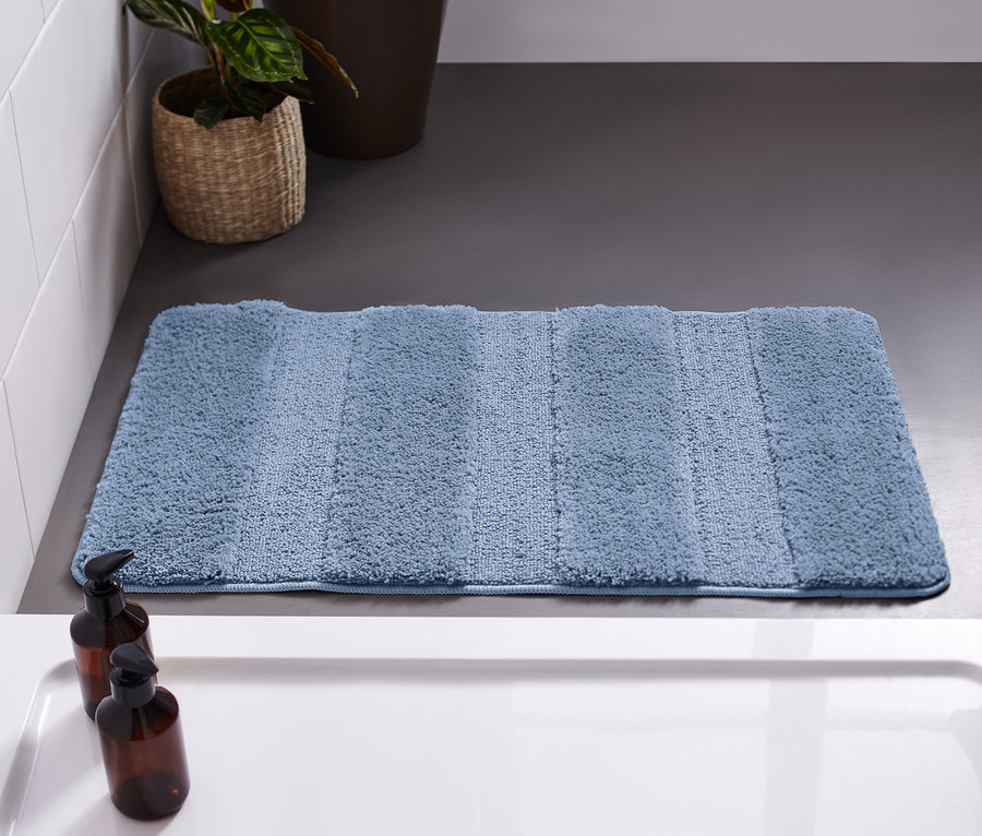 Eine blaue Wellness-Badematte, ca. 50 x 80 cm, liegt auf einem grauen Boden neben einer weißen Badewanne.