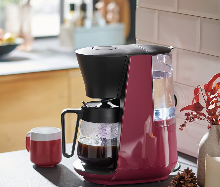 Beerenfarbene »Let's Brew« Filterkaffeemaschine steht neben einer roten Tasse Kaffee auf einer Küchentheke.
