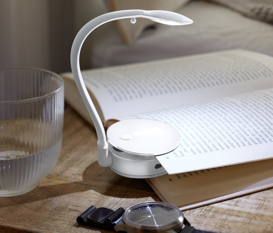 Auf einem Nachttisch liegen ein offenes Buch, eine LED-Leseleuchte, ein Glas Wasser und eine Armbanduhr.